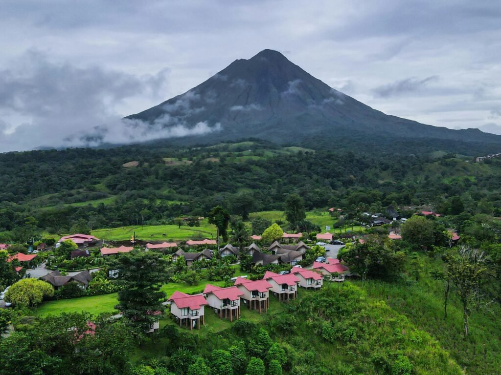 A scenic village with vibrant rooftops nestled below a towering volcano under a cloudy sky.