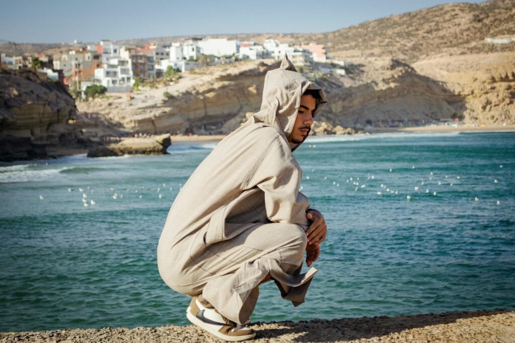 A young man in traditional attire crouches by the sea in Imsouane, Morocco, showcasing the stunning coastal landscape.