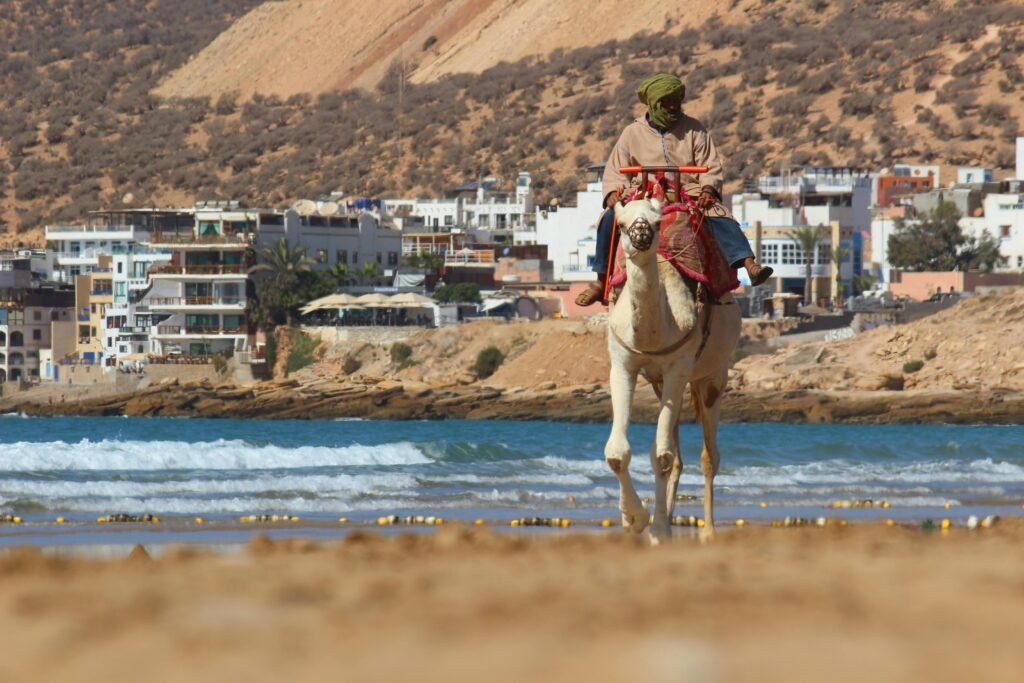 A man riding a camel along the picturesque beach of Taghazout, Morocco with scenic coastal views.